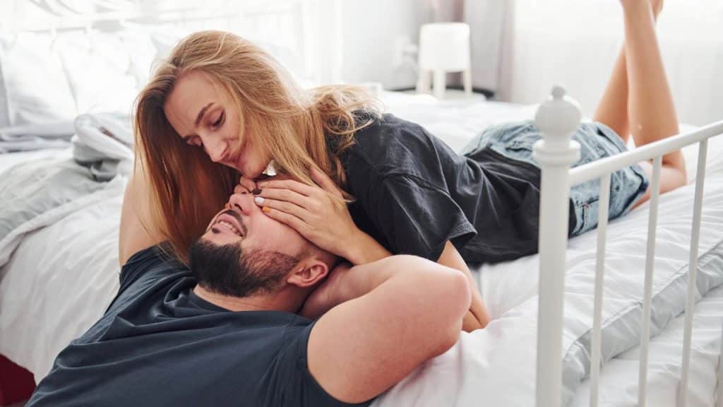 A smiling woman lying on a bed covers a man's eyes with both her hands.