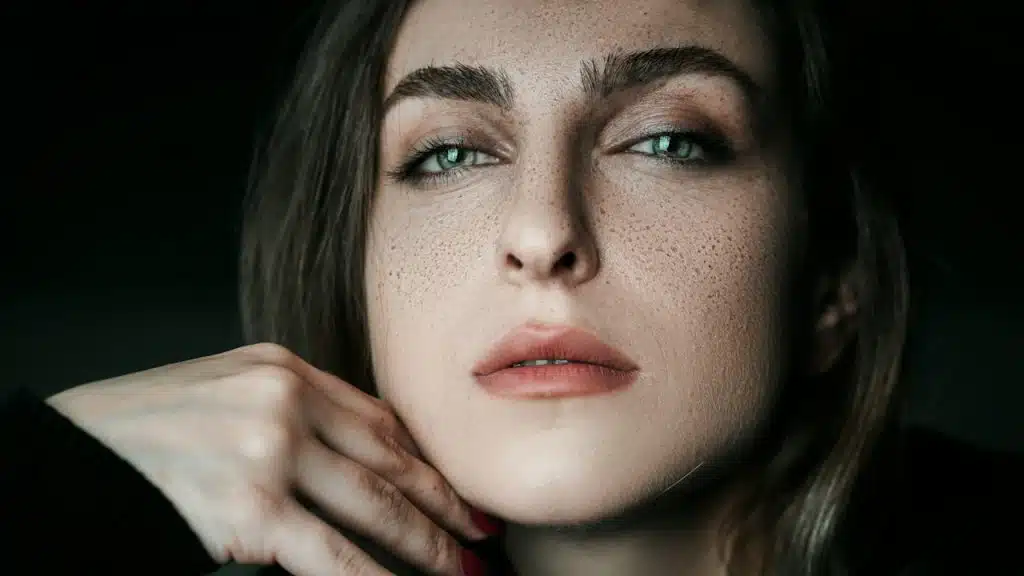 A freckled woman with her hand on her cheek is looking at the camera with half-closed eyes.