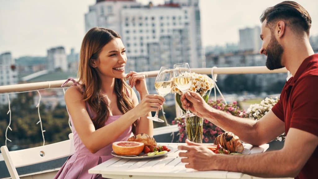 A couple clinking wine glasses during a rooftop meal with a city view.