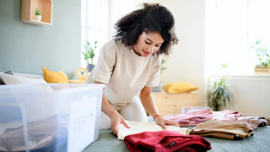 A woman folding clothes on a bed next to a plastic storage bin.
