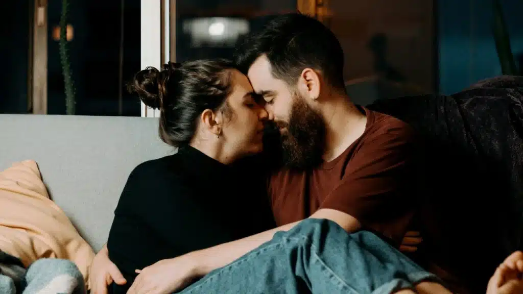 A man and woman sit closely on a couch, touching foreheads with eyes closed.