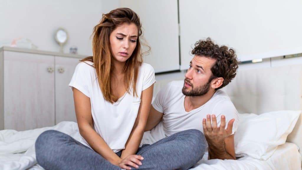 A man gestures with an open hand toward a woman sitting on a bed looking away.