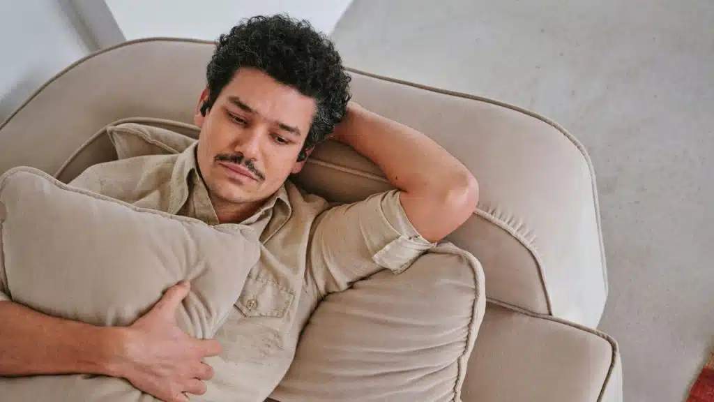 A man with a mustache lies on a tan sofa, resting his head on his hand.