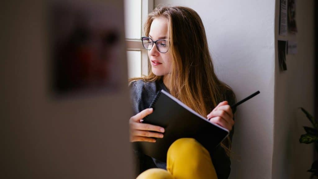 A woman with glasses writes in a notebook while sitting by a window.