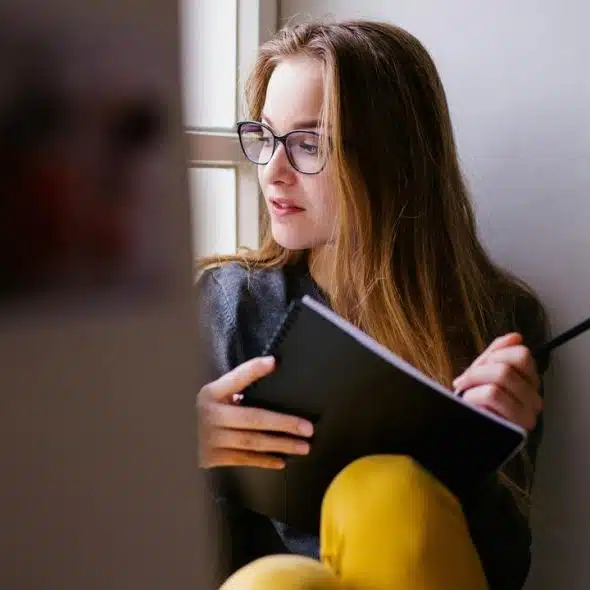 A woman with glasses writes in a notebook while sitting by a window.