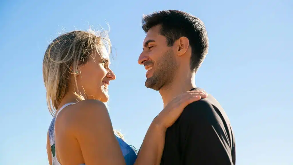 A smiling man and woman stand close together outdoors against a clear blue sky background.