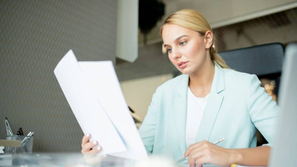 A woman in a light blue blazer holds white papers and a pen at a desk.