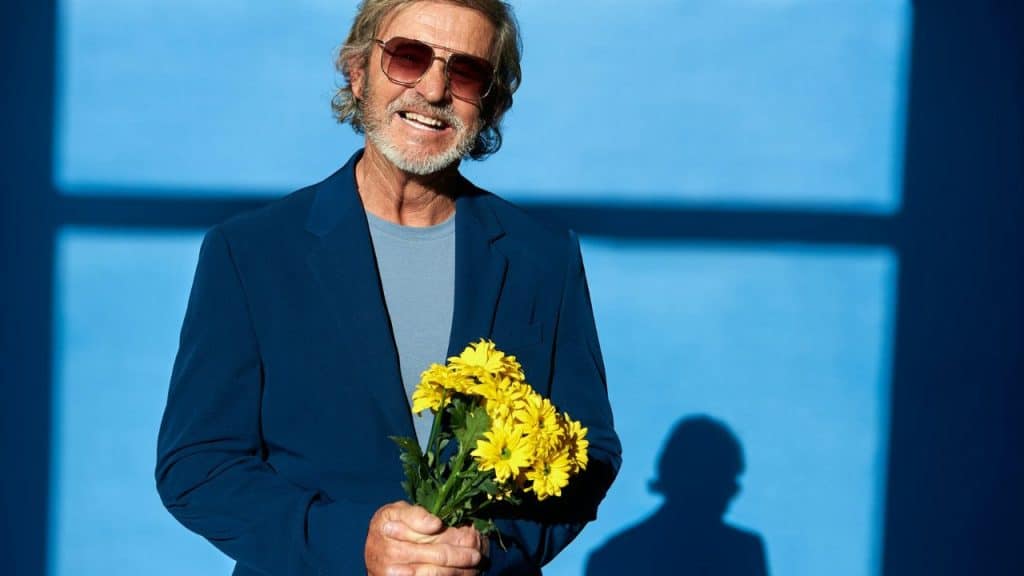 A smiling man with a grey beard wearing sunglasses and a blue suit holding yellow flowers.