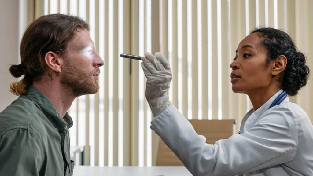 A doctor in a white coat shines a light into a man’s eye during an exam.