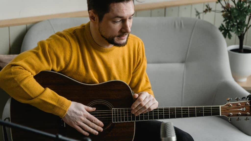 A man in a yellow sweater sits on a couch while holding an acoustic guitar.