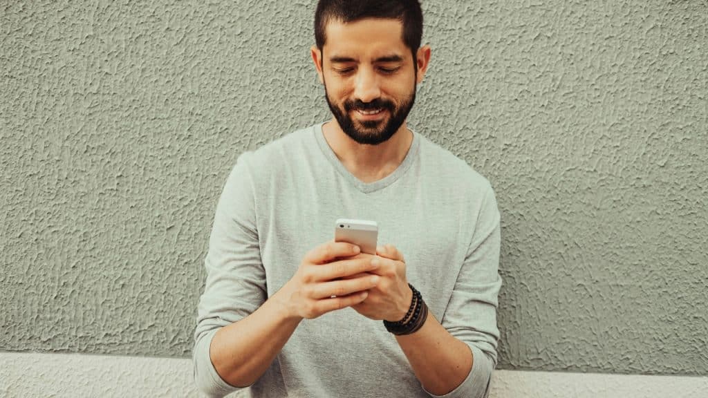 A smiling man with a beard stands against a textured wall while using a smartphone.