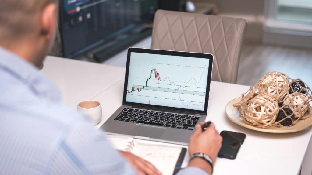 A laptop on a desk displays a financial candlestick chart with green and red bars.