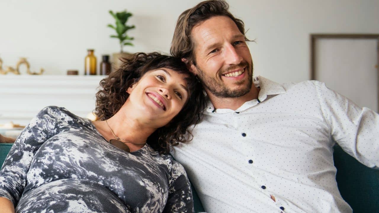 A smiling man and woman sit closely together on a sofa in a bright room.