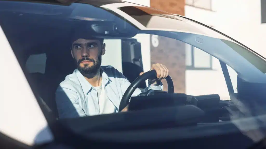 A bearded man with both hands on the steering wheel looks out a car window.