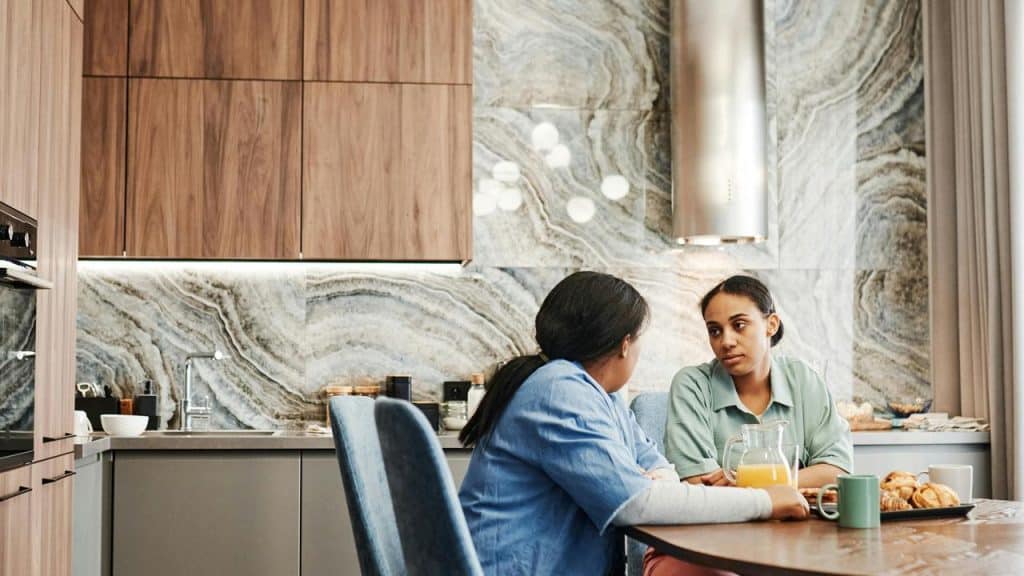 A woman talking with another woman at a kitchen table with breakfast items.
