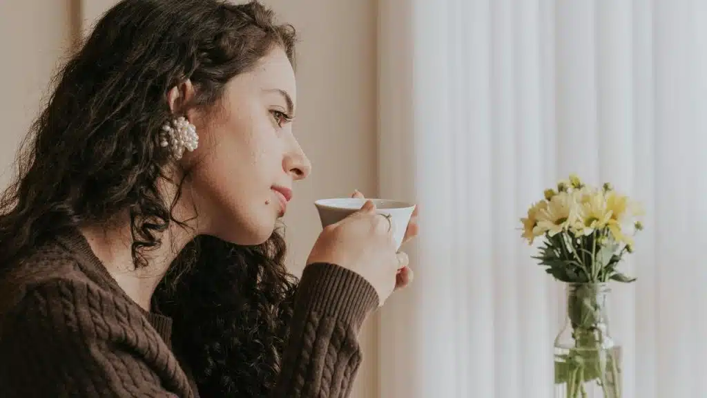 A woman sitting at a table, holding a cup and looking thoughtfully at flowers.