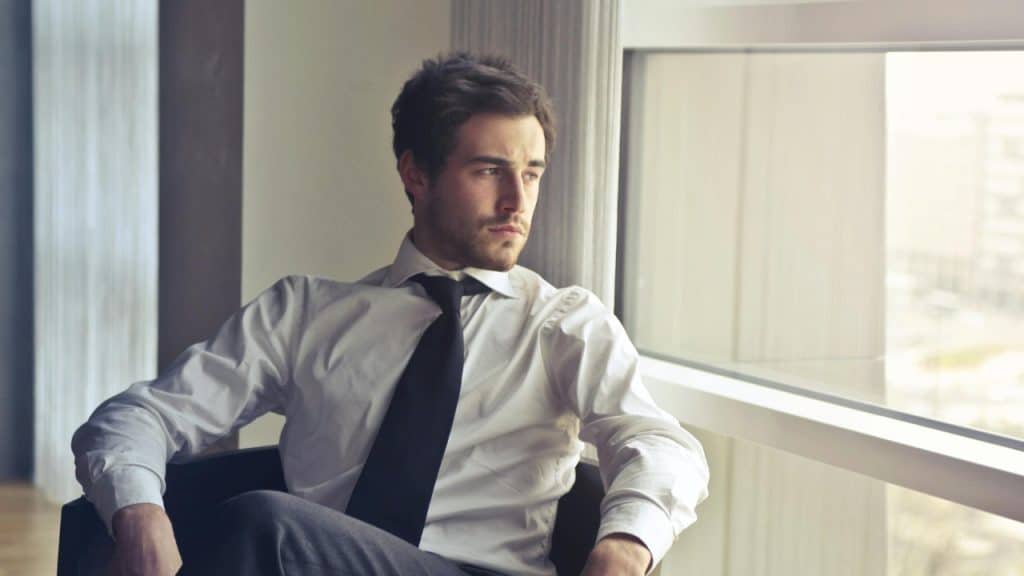 A man wearing a white shirt and black tie sits in a chair near a window.