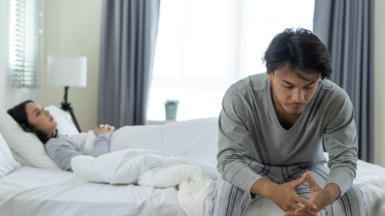 A man sitting on the bed looking anxious and clasping his hands while a woman lies in the background.