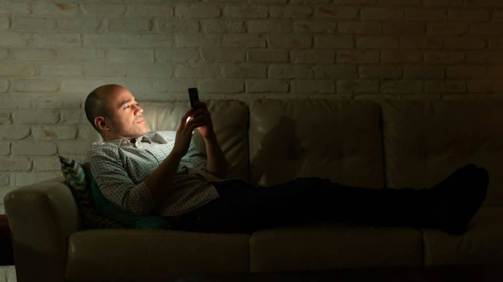 A man lies on a couch in a dark room while looking at a glowing smartphone screen.