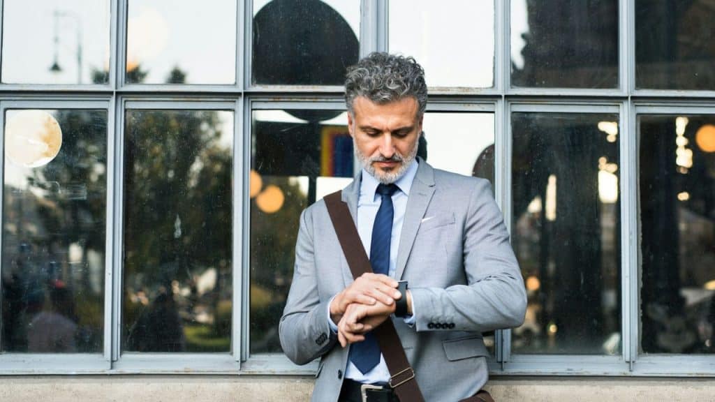 A man in a grey suit leans against a window and looks at his watch.