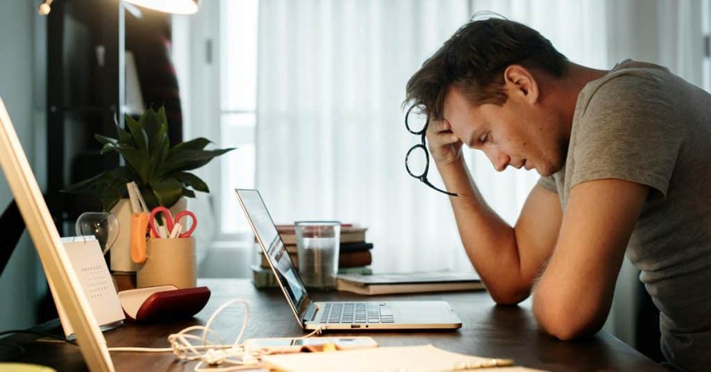 A person sits at a desk holding glasses while looking at a laptop.