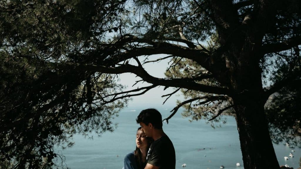 A couple standing under large tree branches with the sea in the background.