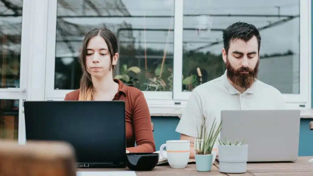 A woman and a bearded man sit side-by-side outdoors while working on separate laptops.