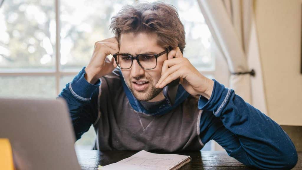 A man wearing glasses talking on the phone looking frustrated at his laptop.