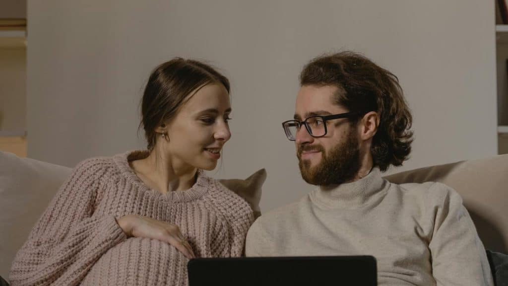 A couple sitting on a couch looking at each other with a laptop in front of them.