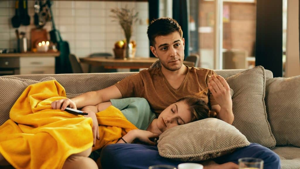 A man watches television while a woman sleeps with her head resting on his lap.