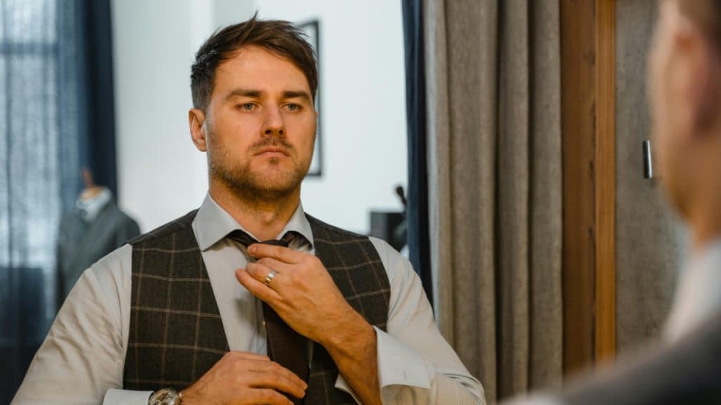 A man in a vest and tie adjusts his necktie while looking into a large mirror.