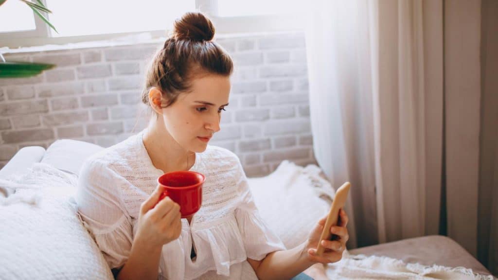 A woman holding a red mug while looking at her phone on a couch.