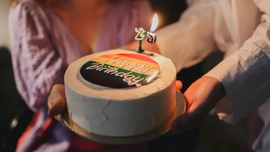Hands hold a white cake with a "Happy Birthday" topper and a lit candle.