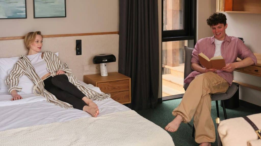 A woman relaxes on a bed while a man sits in a chair nearby reading a book.