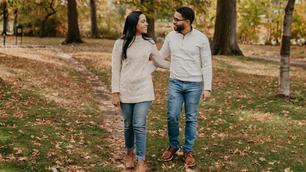 A smiling man and woman walk together through a park covered in fallen autumn leaves.