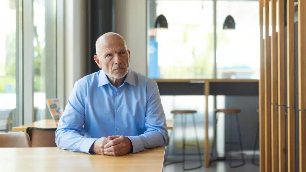 A bald man in a light blue button-down shirt sits at a wooden table indoors.