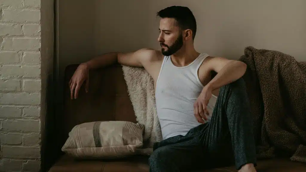 A bearded man wearing a white tank top sits on a brown leather couch indoors.