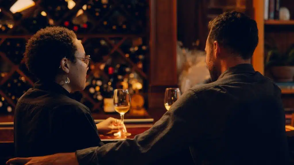 A man and woman sit at a bar with wine glasses in front of shelves.