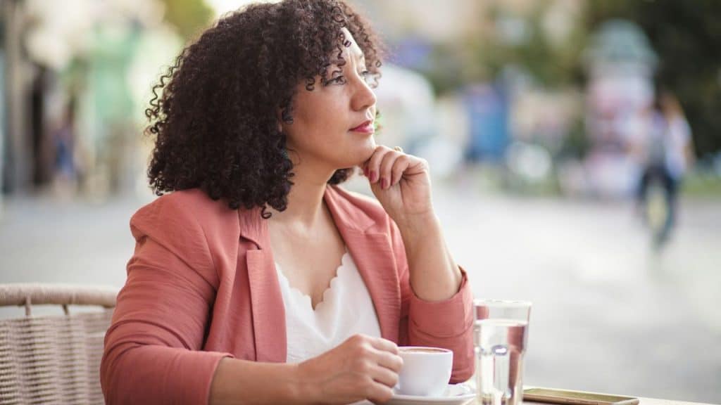 A woman with curly hair sits at an outdoor table holding a white coffee cup.