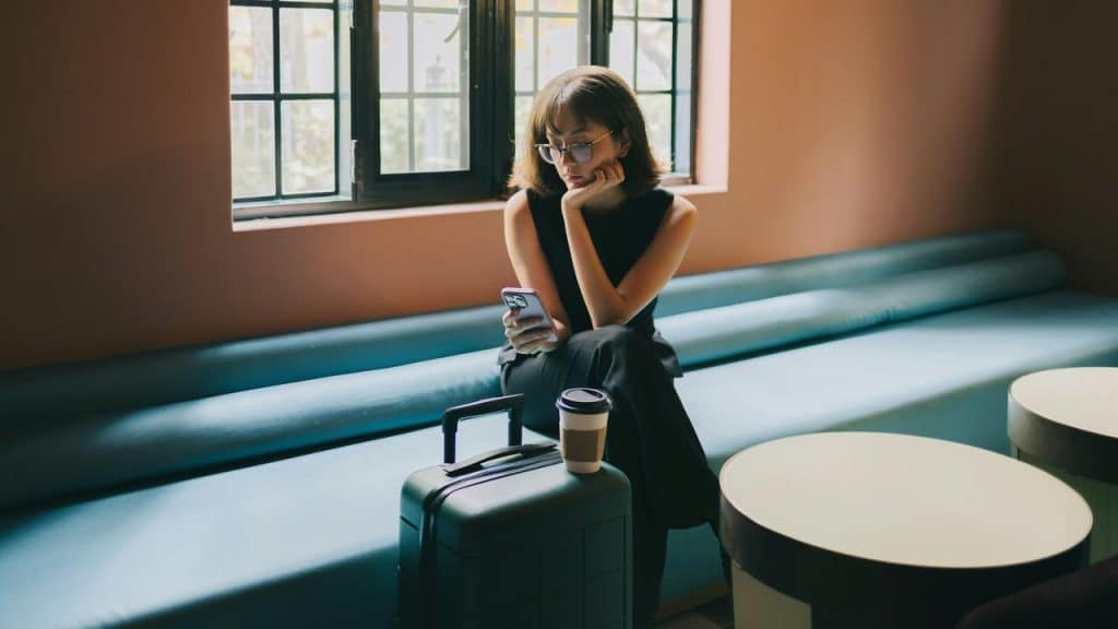 A woman with glasses sitting on a bench, checking her phone beside a suitcase.