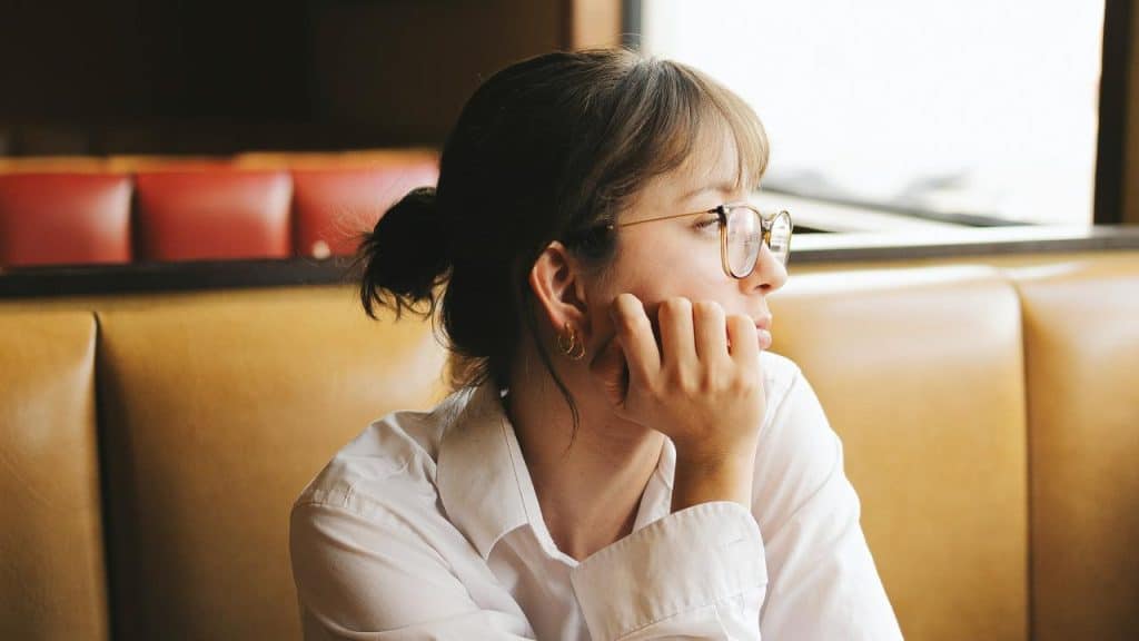A woman wearing glasses sits in a booth, resting her chin on her hand.