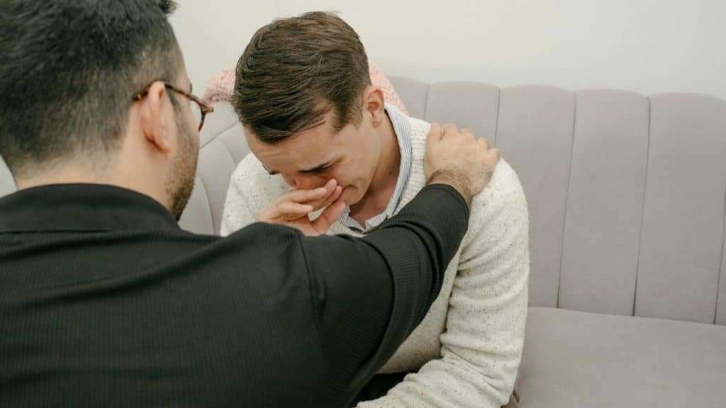 A man comforting a crying friend sitting on a couch.