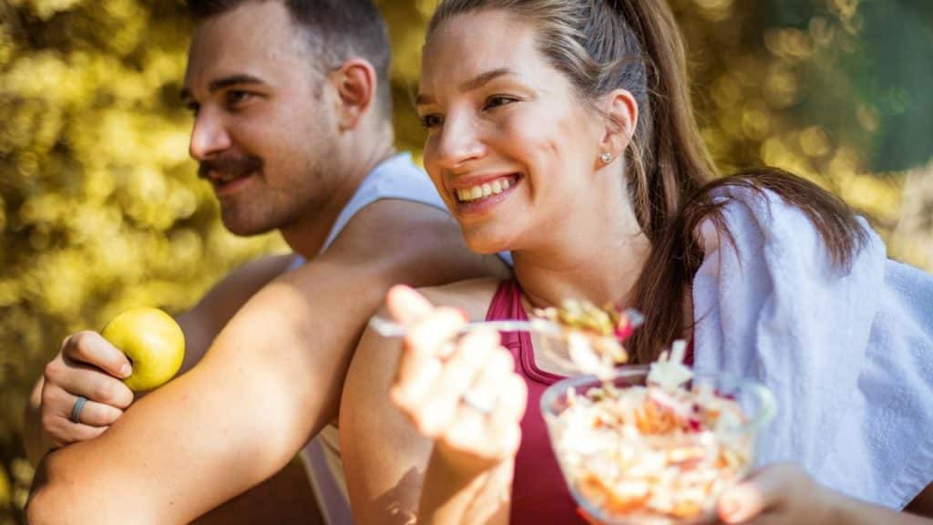 A smiling couple relaxes outdoors, with the woman eating salad and the man holding an apple.
