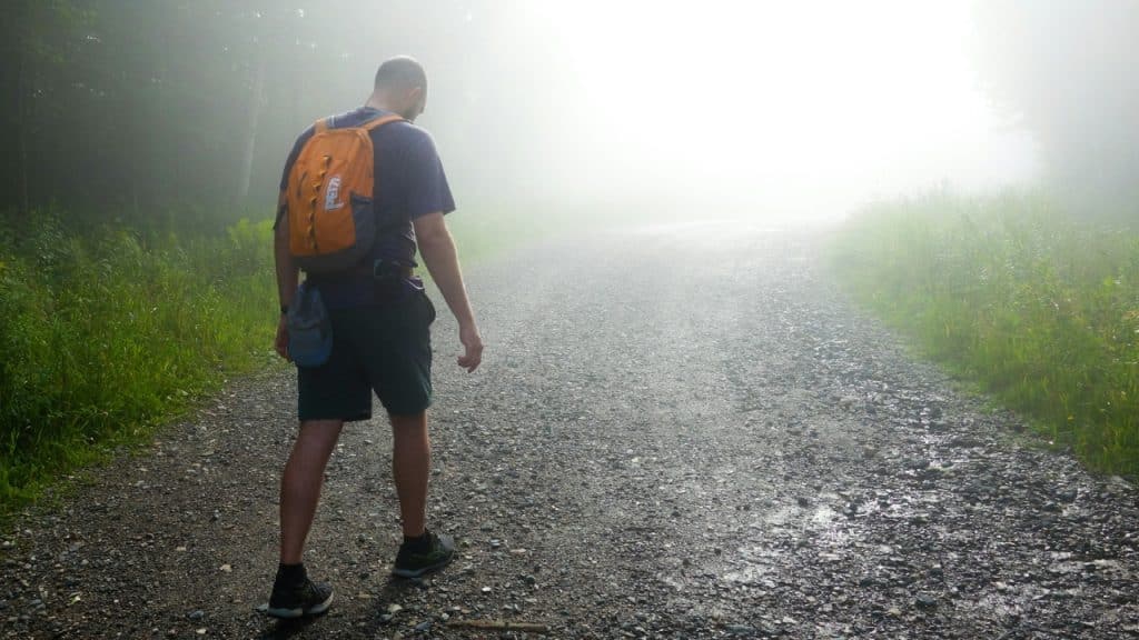A man with an orange backpack walks down a gravel road into thick white fog.