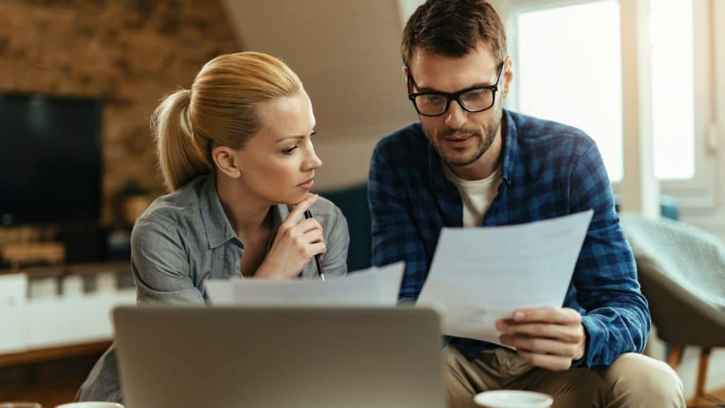 A man and woman sit in front of a laptop while looking at white papers.