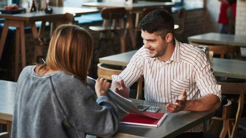 A man in a striped shirt gestures while talking to a woman at a table.