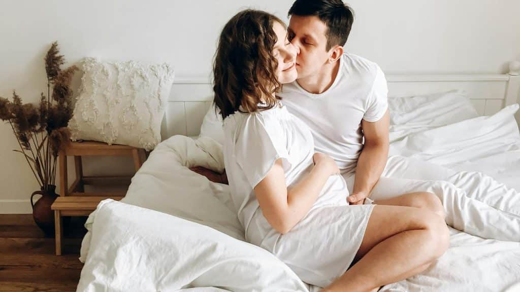 A couple sits on a bed while the man kisses the woman’s cheek.