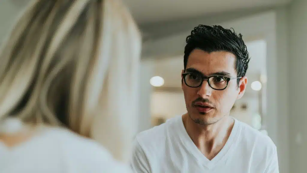 A man wearing glasses looks intently at a woman during a serious conversation in a bright indoor setting.