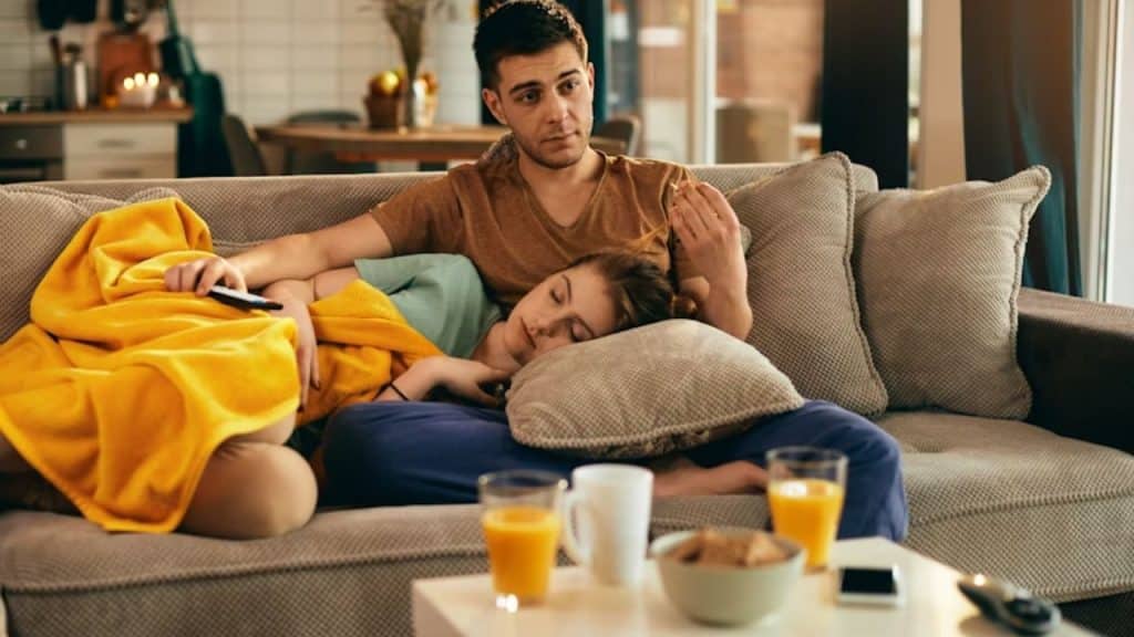 A man is watching TV while a woman lies in his lap and sleeps.