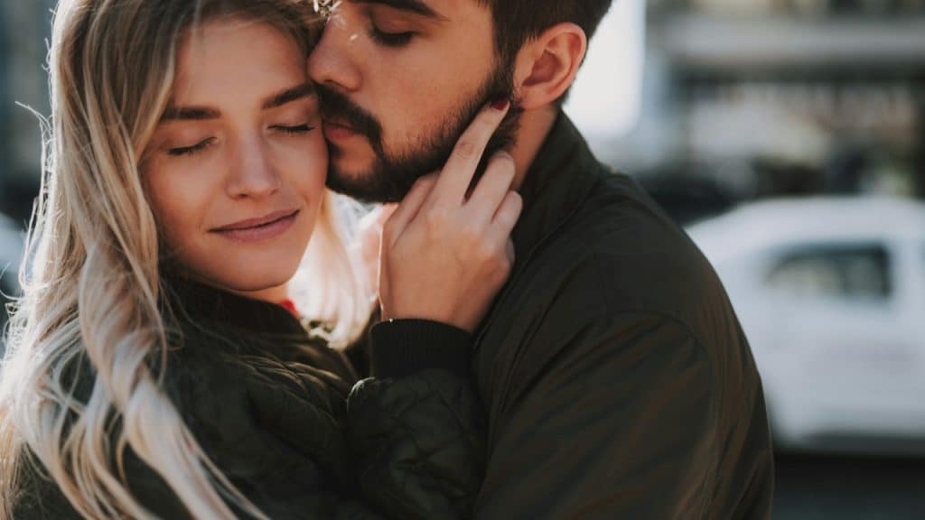 A man leans in to kiss a woman's cheek while she smiles with her eyes closed.